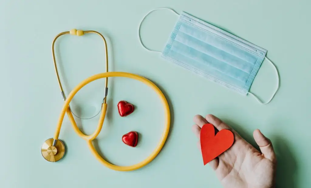 A yellow stethoscope, red hearts, and a mask rest beside a caring hand—symbols of health and community support.