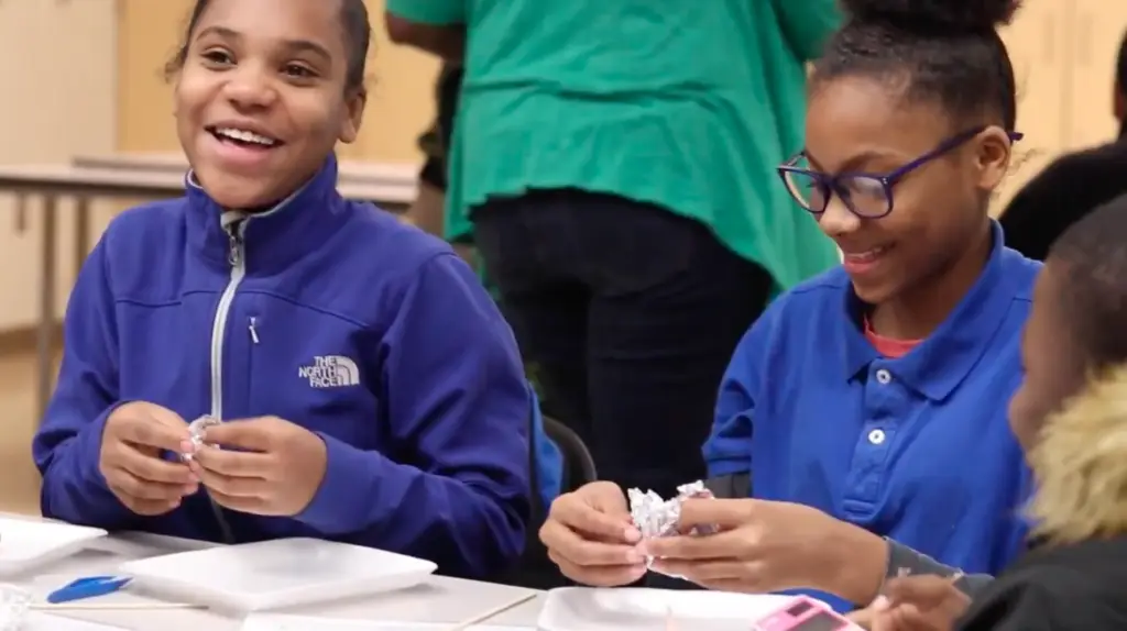 Two girls happily work together at a table, folding foil in a group setting that fosters learning and community.