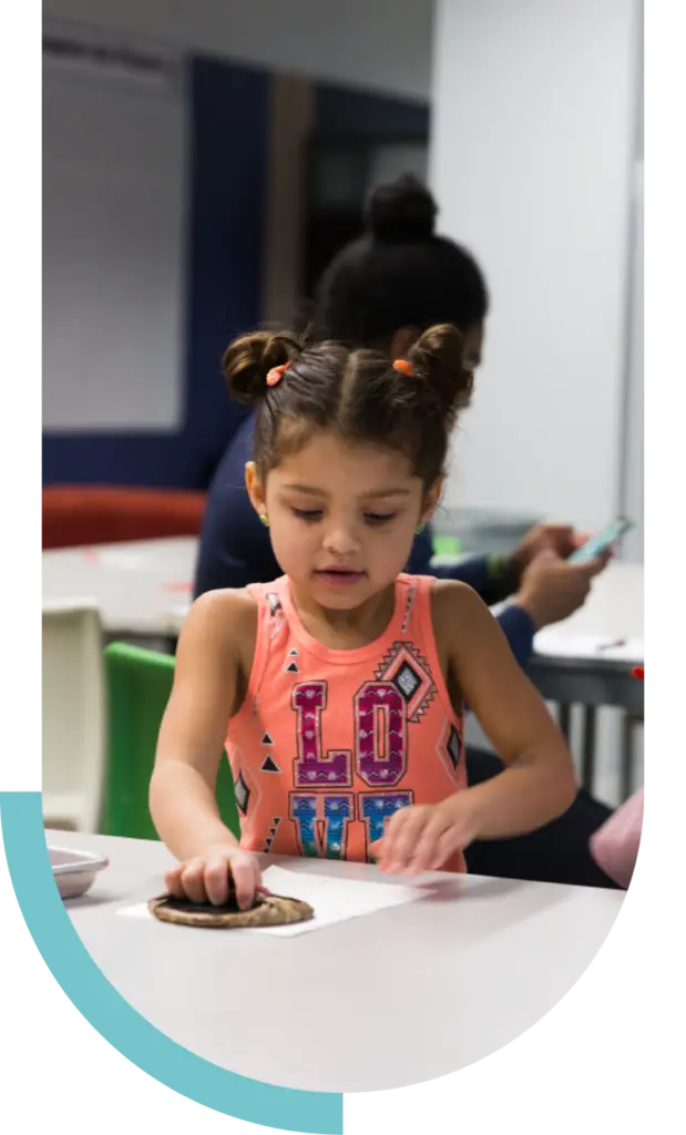 A young girl concentrates on a hands-on activity at a table, while an adult supports learning and growth in the background.