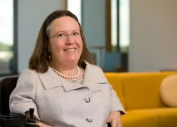 A smiling woman in a wheelchair, wearing glasses and a pearl necklace, brings warmth to an inviting, accessible office space.