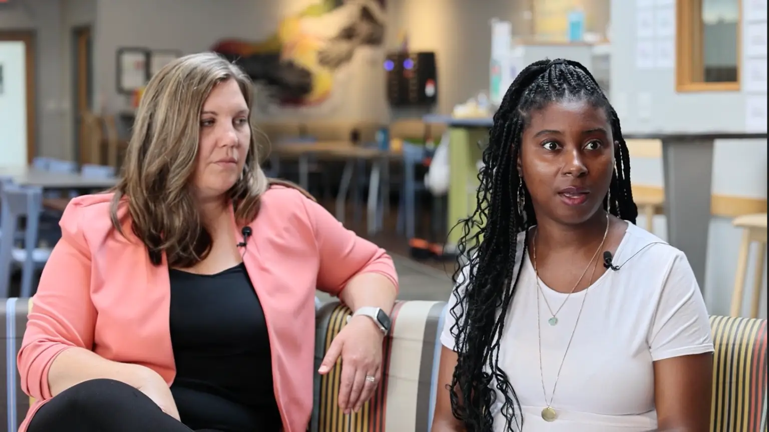 Two women share ideas on a couch in a bright space, highlighting community and open dialogue with clip-on mics.