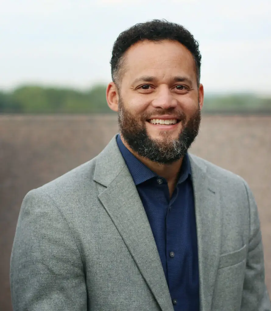 A smiling man in a grey jacket and navy shirt stands outdoors, reflecting warmth and community connection.
