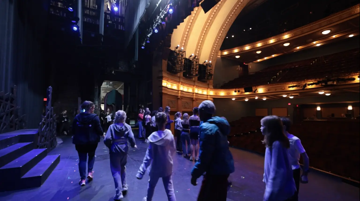 Children and adults gather on a lit theater stage, exploring arts together in an inviting, grand community space.