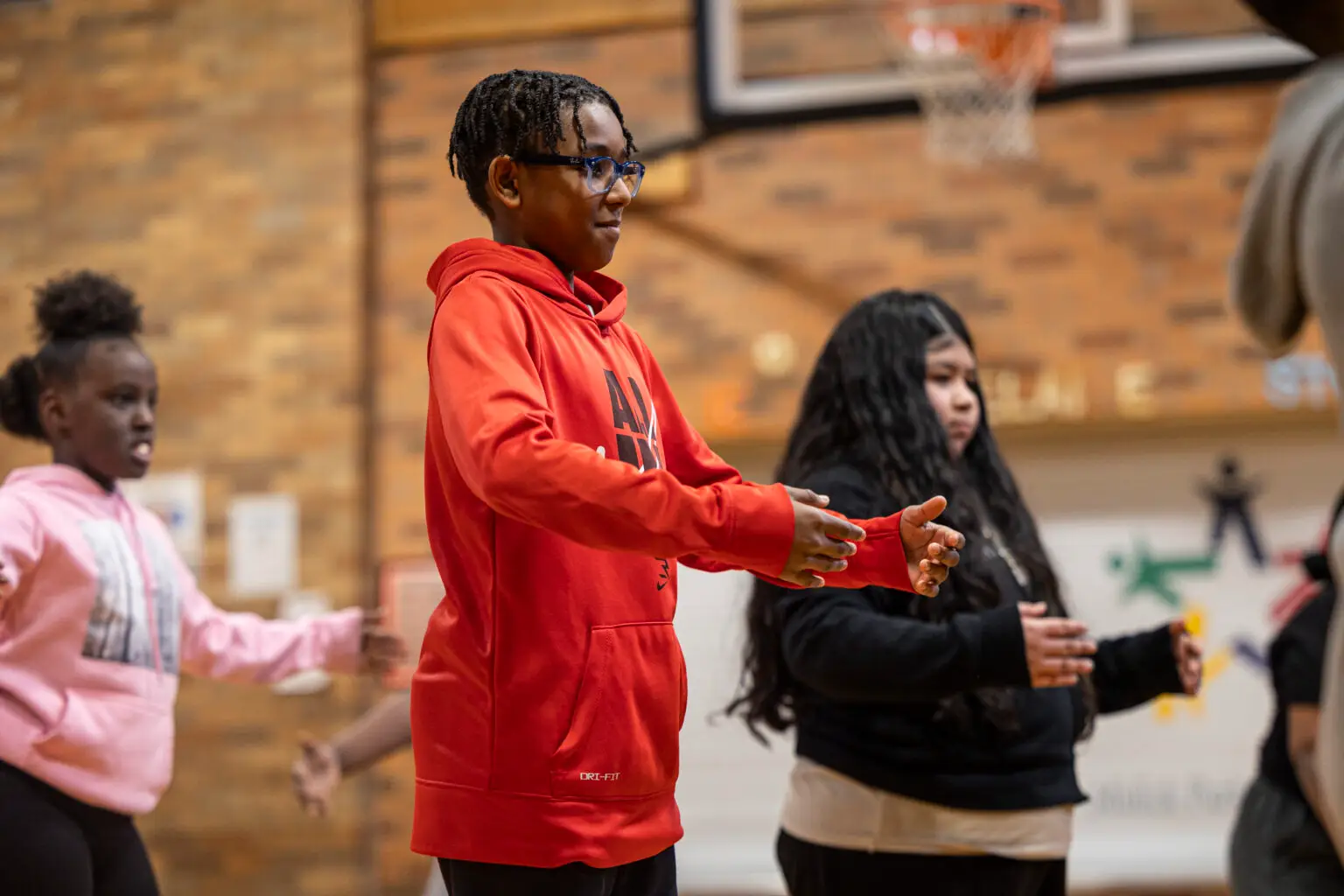 Children participate in indoor activities, raising their arms together. A boy in a red hoodie leads near the basketball hoop.