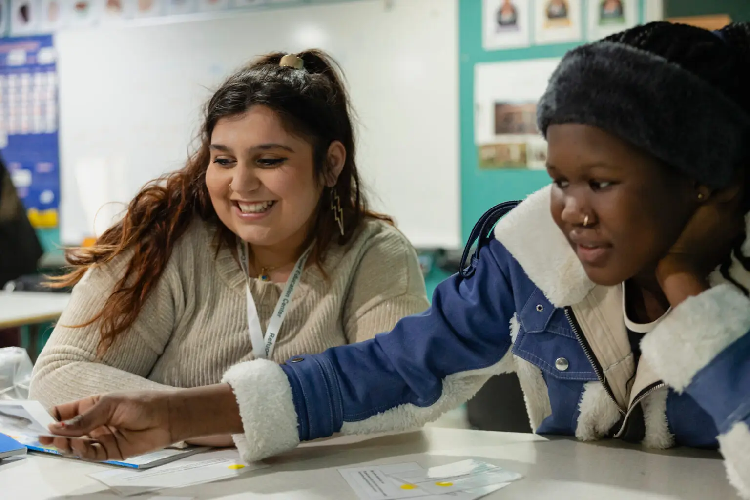 Two young women share a classroom activity, smiling as they collaborate with papers and cards, fostering learning together.