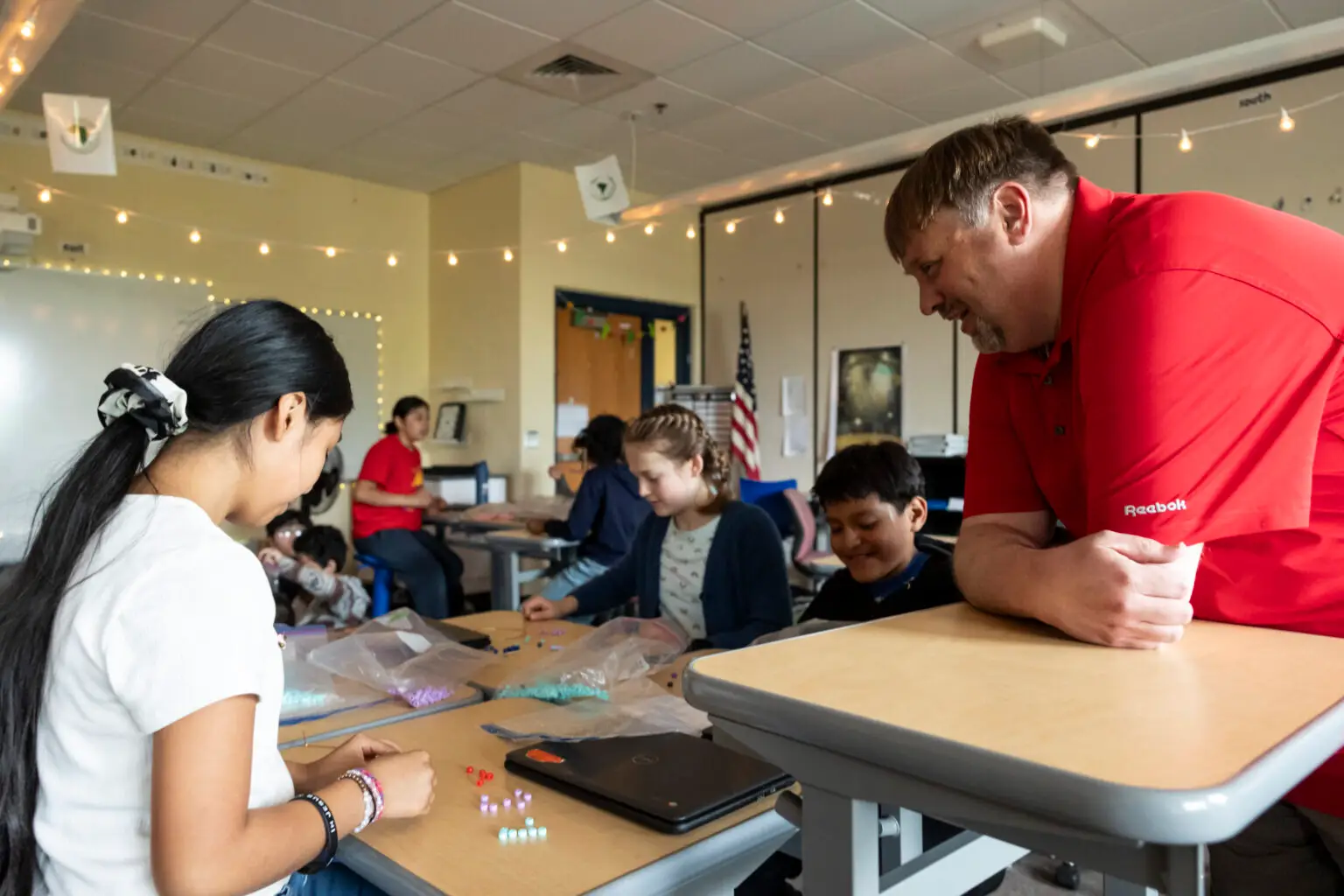 A teacher supports students crafting with beads in a welcoming, well-lit classroom, fostering creativity and community learning.