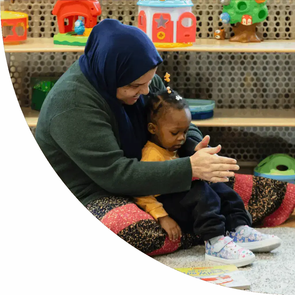 A caring adult helps a young child clap hands together in a vibrant playroom, fostering learning and joyful connection.