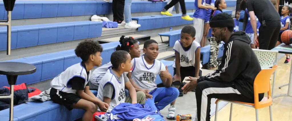 A coach engages young All Stars players in a gym, fostering teamwork and learning as others look on.