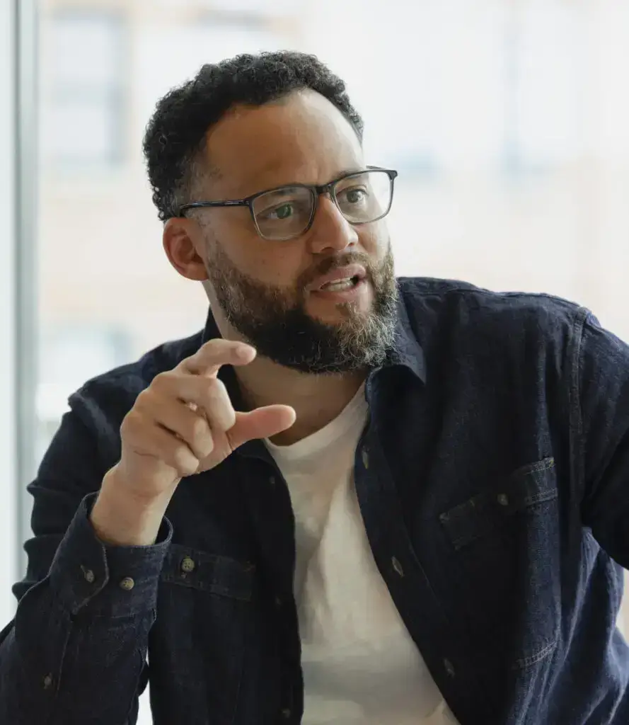 A bearded man in glasses gestures as he speaks, engaging others in a thoughtful, community-focused discussion indoors.