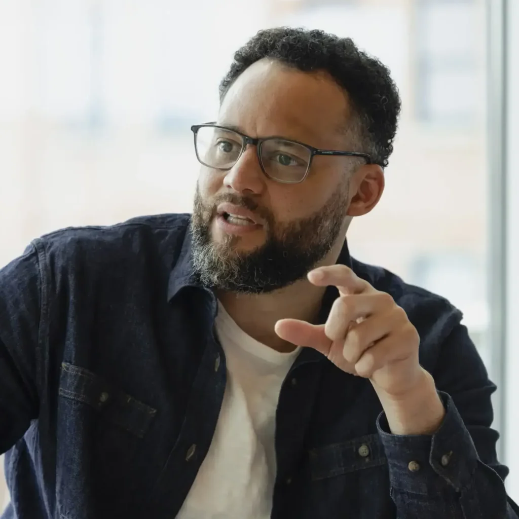 A bearded man in glasses gestures as he speaks, engaging others in a thoughtful, community-focused discussion indoors.