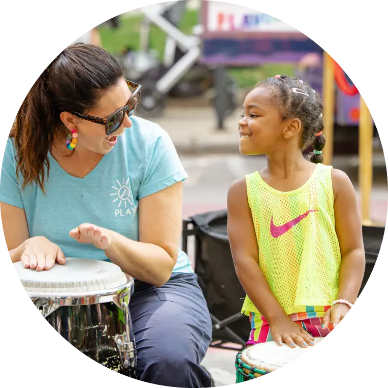 A woman and young girl share a joyful moment drumming together outside, highlighting connection and community.