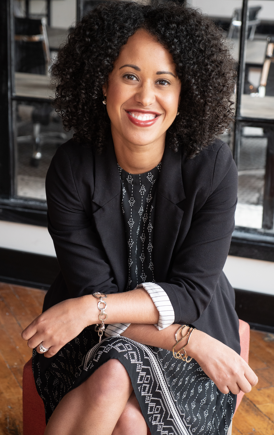 Smiling woman in a blazer and patterned dress sits confidently in a modern office, embodying leadership and opportunity.