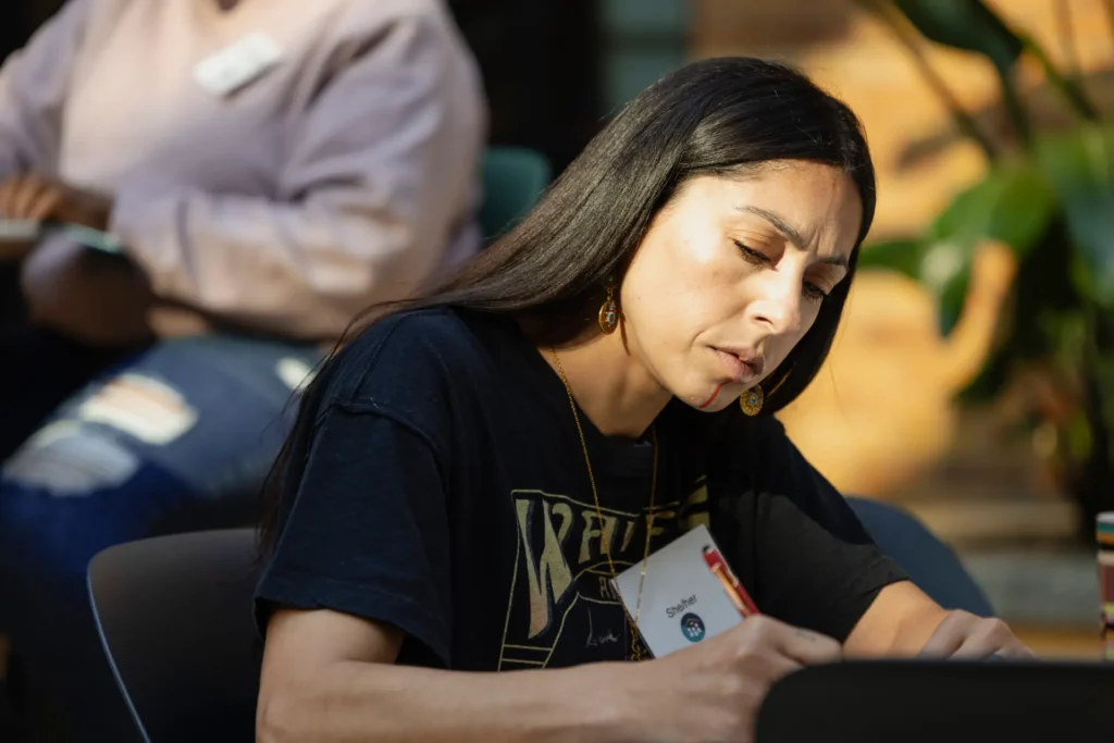 A woman with long dark hair sits at a table, focused on writing in a notebook. She wears a black T-shirt, earrings, and a name badge. Another person is blurred in the background. Sunlight highlights her face.