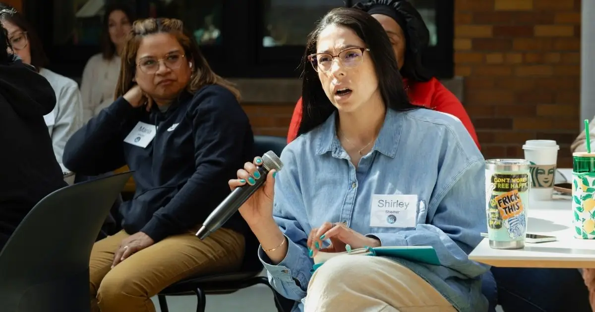 A woman holding a microphone and speaking at an indoor event, with a name tag reading Shirley. She is seated next to others, with drinks and papers on the table in front of her.