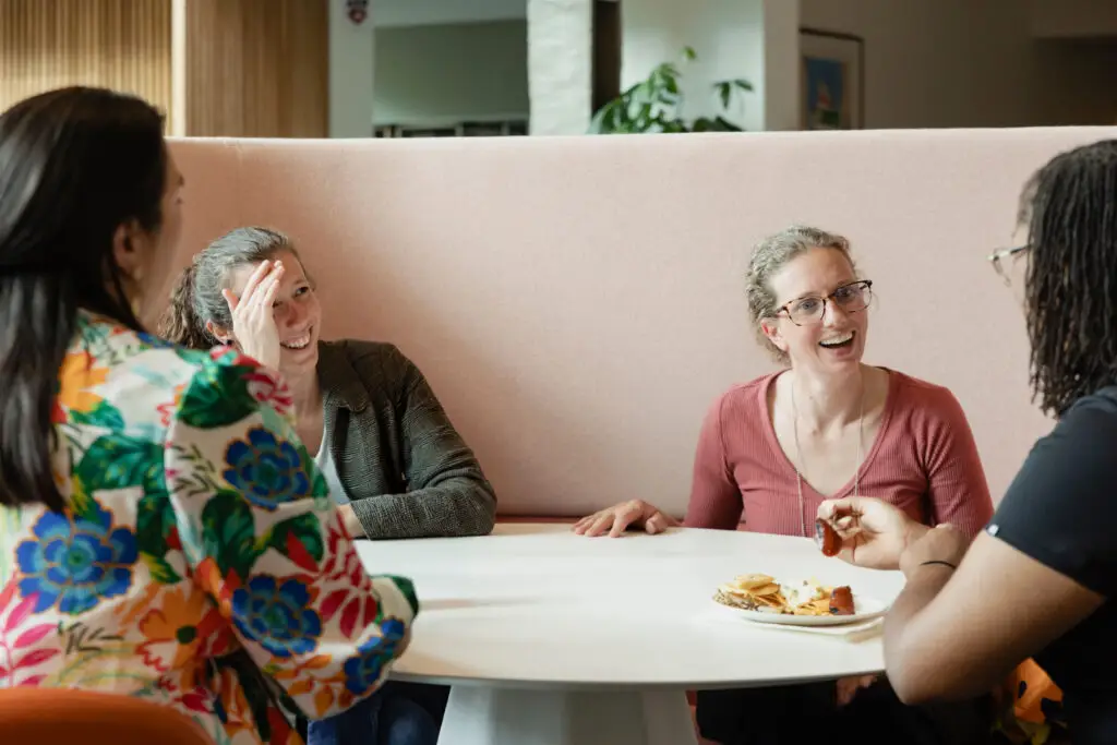 Four people sit around a white table, talking and laughing. Two women face the camera, one wearing glasses, while another gestures with her hand. Food is on the table in front of them. The setting appears casual and friendly.