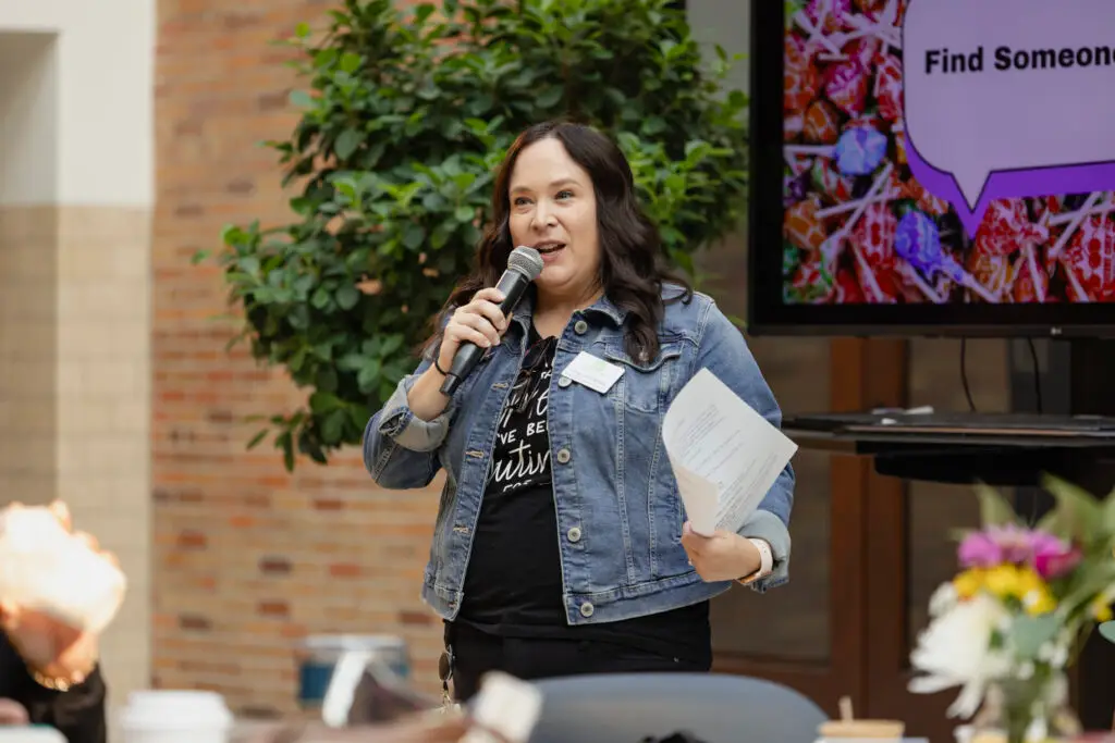 A woman with long dark hair, wearing a denim jacket, holds a microphone and papers while speaking. She stands indoors near a large leafy plant, with a screen and flowers visible in the background.