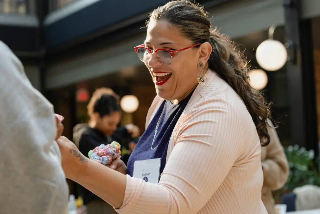 A woman with red glasses and long hair smiles brightly while handing out colorful candies at an indoor event. She wears a light pink jacket, earrings, and a name tag, and appears to be enjoying the lively atmosphere.