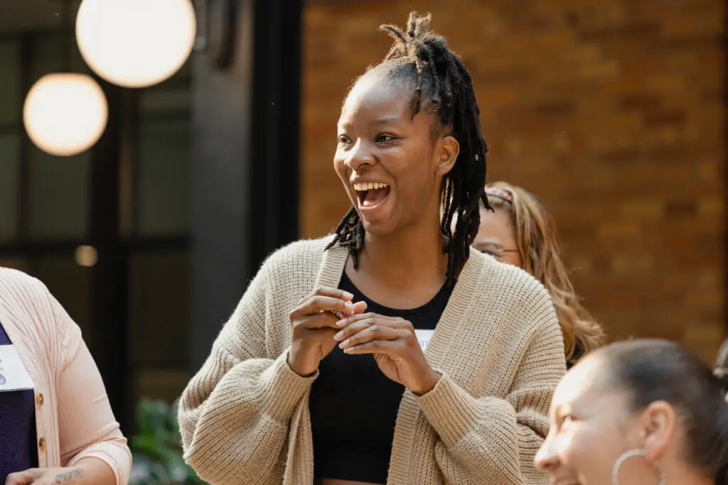 A woman with braided hair smiles and laughs while standing indoors, wearing a beige cardigan and black top. Other people are nearby, and warm lighting and a brick wall are visible in the background.
