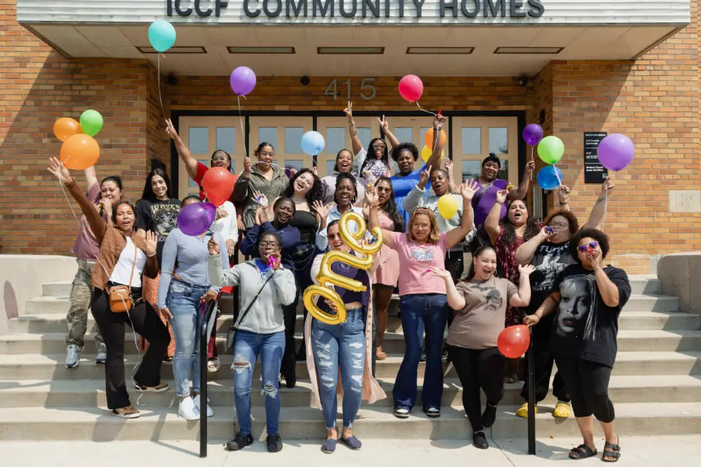 A diverse group of people stand on building steps, cheering and holding colorful balloons. Two people at the front hold gold letter balloons spelling one. The building sign reads ICCF Community Homes.