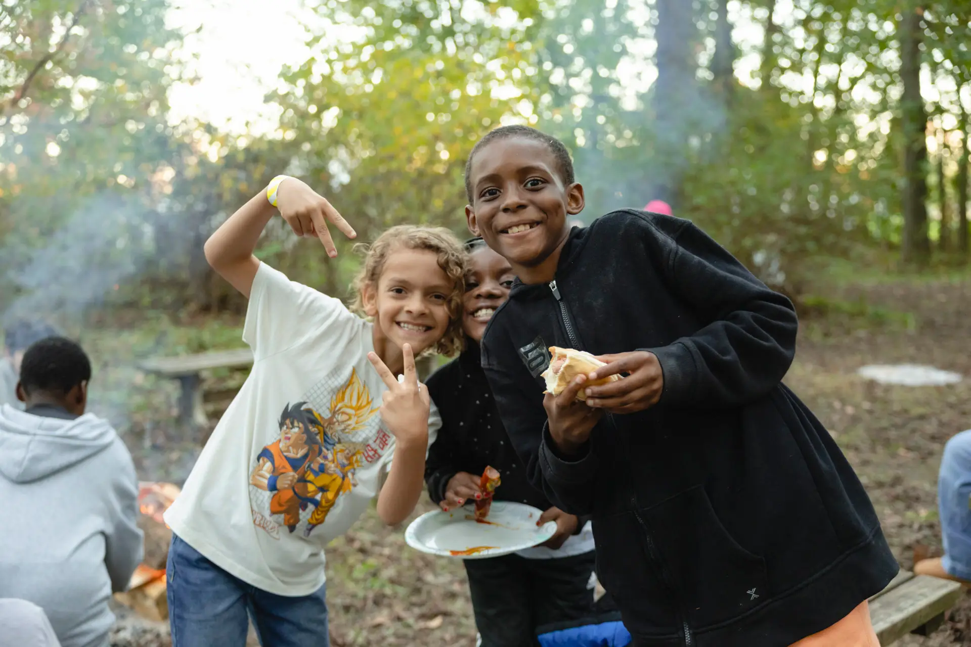 Two smiling children pose outdoors near a campfire, one holding food and the other making a peace sign. Other kids sit in the background among trees and smoke from the fire. The scene suggests a fun camping activity.