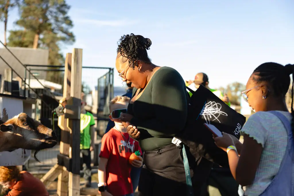 A woman and a girl interact with a goat at an outdoor petting zoo on a sunny day, while other children and adults are visible in the background.